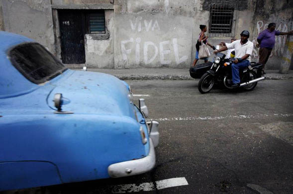 A vintage car passes by a wall with a political slogan that reads 'Viva Fidel'