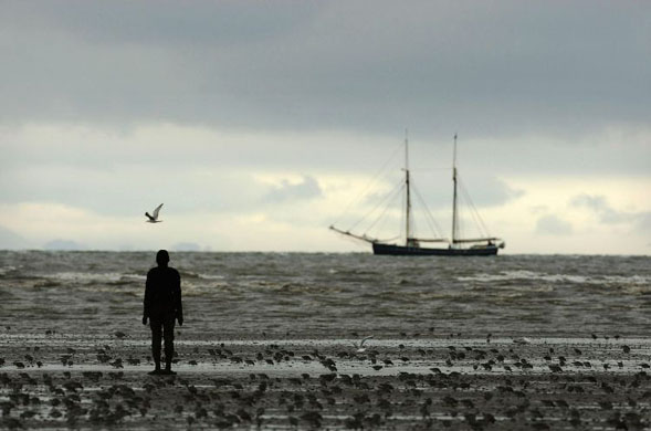 Liverpool, UK: A figure from Antony Gormley's 'Another Place'. One of the Tall Ships can be seen in the background