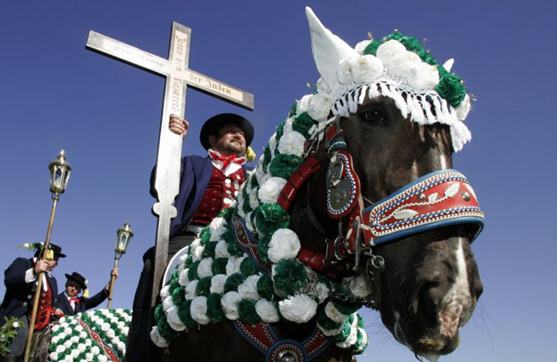 A man dressed in traditional Bavarian costume rides his horse and holds a wooden cross