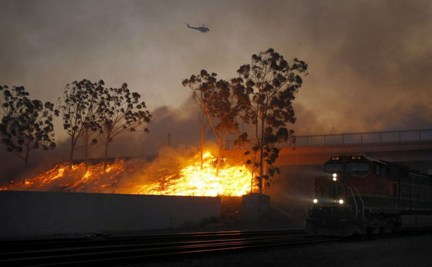 A train moves to get past a wind driven wildfire as a fire helicopter circles overhead in Yorba Linda, California