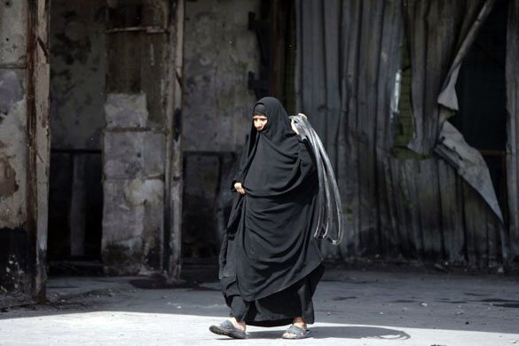 Baghdad, Iraq: A woman carries rubber pipes as she walks by the site of a past explosion at Abu Dshirin market