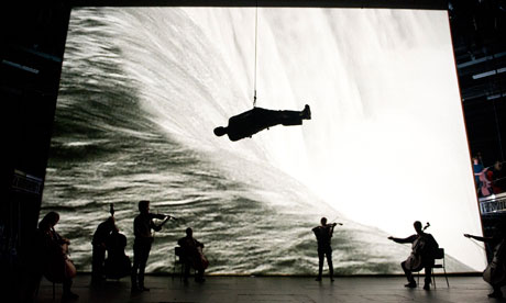 Survivor: string musicians against a huge video screen with a man suspended horizontally overhead