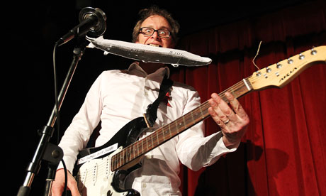 Simon Munnery with a guitar gripping a cardboard airship in his teeth