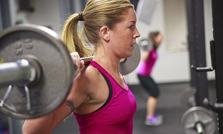 Blond women in pink top doing back squats with barbell in weight gym.