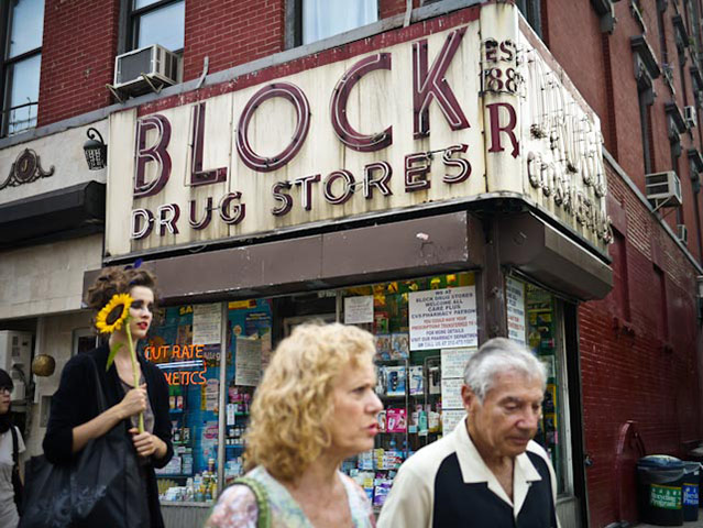 Panasonic G3 | NYC: Three people walk past a drug store in daytime NYC, one holds a sunflower