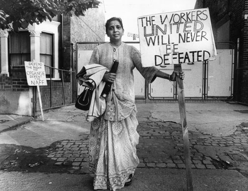 Jayaben Desai: A woman stands to camera alone holding handbag, coat and placard