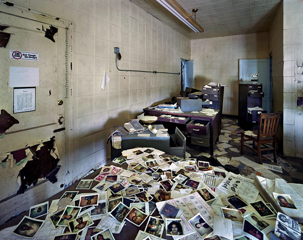 Photos from police dossiers litter the floor of a police office.  Filing cabinets have their draws left open and the paint on a secure room door peels