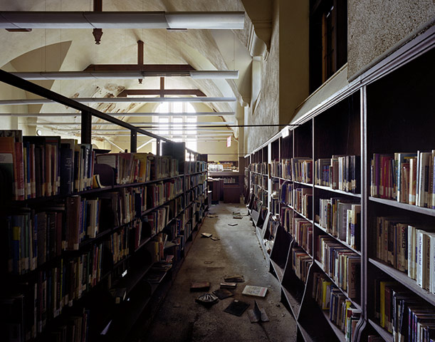 A photo of a library in disrepair.  Books line shelves or are on the ground, light filters through a dirty window and the paint is peeling from the ceiling