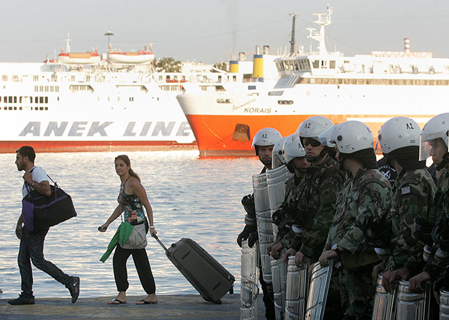Business Pix of Week: Tourists carry their luggages past riot policemen in the port of Piraeus