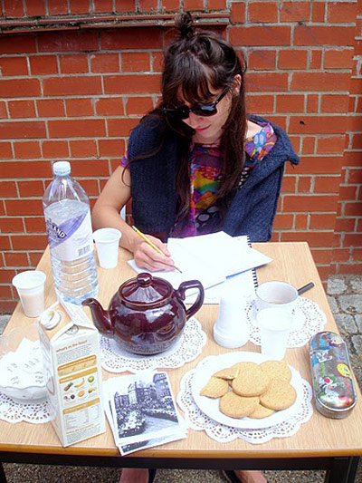 Liverpool Road: A young woman writes notes at a table laden with tea & biscuits