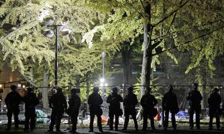 Police in riot gear silhouetted against barbed wire fencing ...