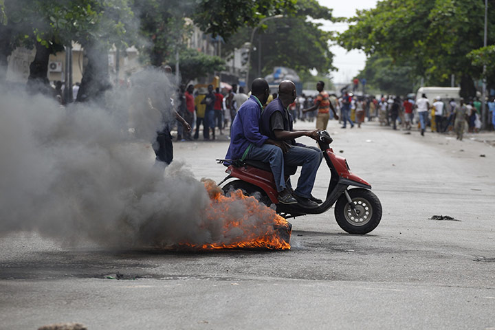Ivory Coast violence: Men ride a moped past burning debris shortly after civilians were shot at