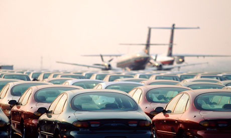 Cars parked in an airport carpark. In the distance two planes sit on the runway.