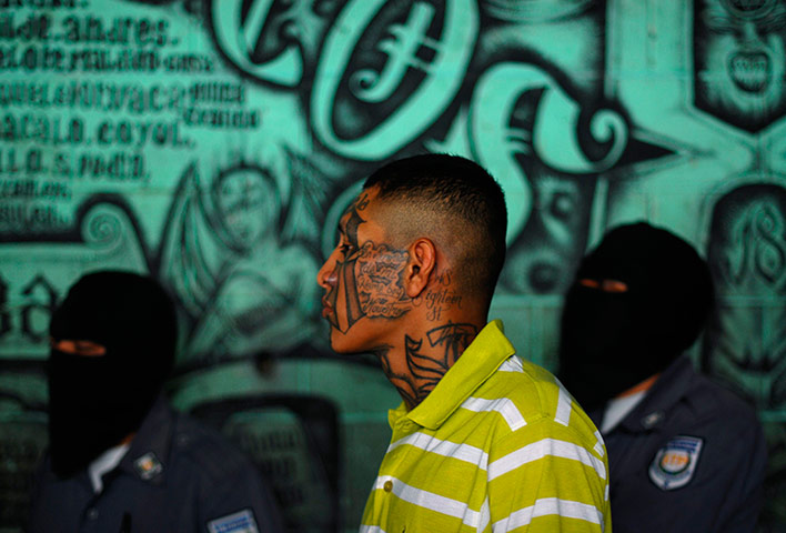 Longer View: A gang member and inmate stands near masked police officers 
