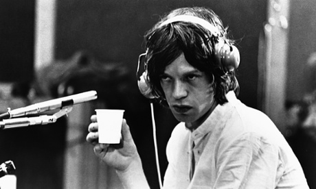 Mick Jagger holds a cup and wears headphones.