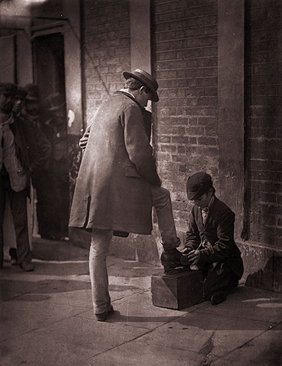 Street Life in London: A young independent shoeblack shines the boots of a city gent