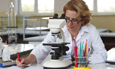 Female researcher taking notices while she is using a microscope.