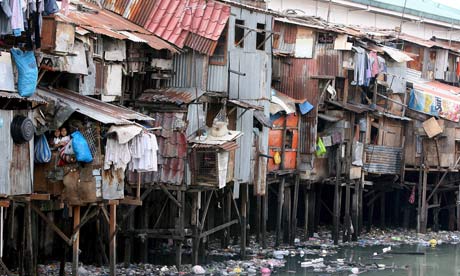 A row of shanties hangs precariously over a filthy river full of rubbish