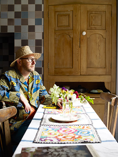 Homes - Hollicks house: Man dressed in blue and yellow jacket and straw hat sitting in kitchen