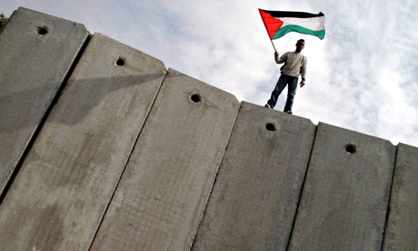 A man stands on the wall along the border with Israel and waves a Palestinian flag