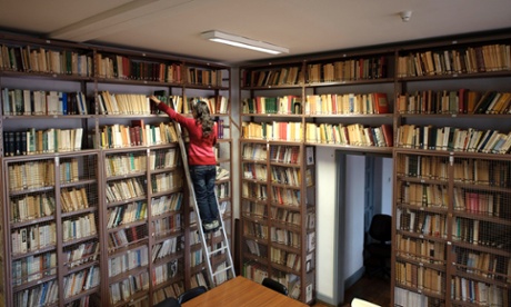 Wide angle view of young woman on metal ladder searching for books on a library ladder.