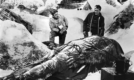 Still from a black and white movie; two men in climbing gear stand in a snowy mountain scene, looking down at a giant, hairy, bipedal animal covered in a tarpaulin and strapped to a stretcher.