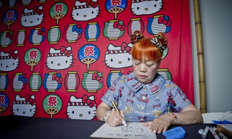 Official illustrator Yuko Yamaguchi sits and draws, an array of Hello Kitty designs behind her