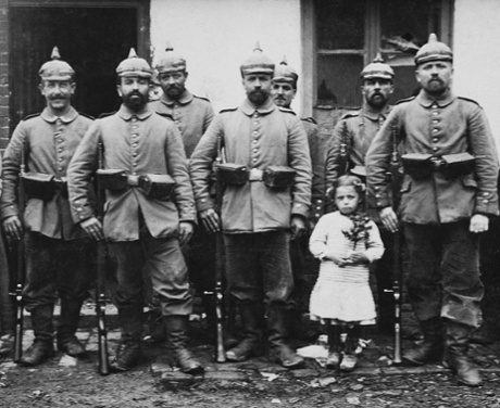 A little girl clutches a small Christmas tree while standing with a German military outfit early in WWI somewhere in war torn France.