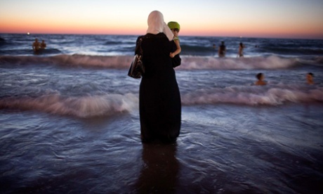 A Palestinian woman dressed from head to toe stands in the sea holding her baby as others swim