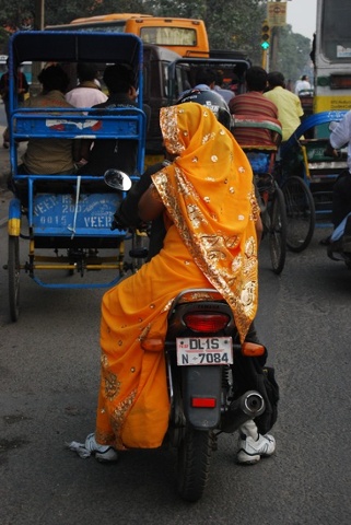 A woman in an orange sari sits astride a motorcycle on the streets of Old Delhi.