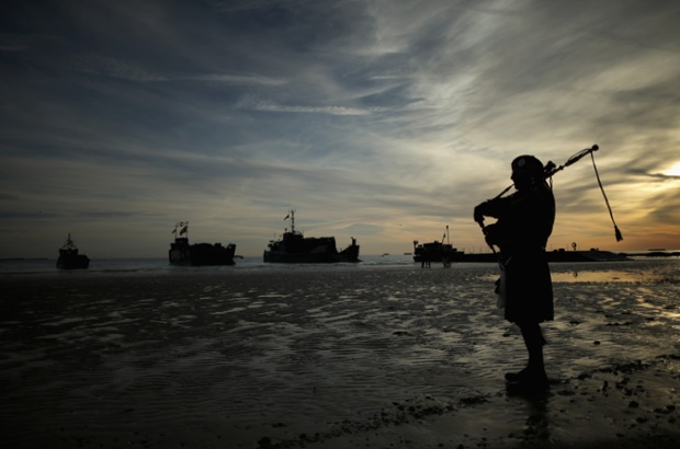 A piper plays a lament on Gold Beach as landing craft from the Royal Marines arrive at Arromanches Les Bains, France.