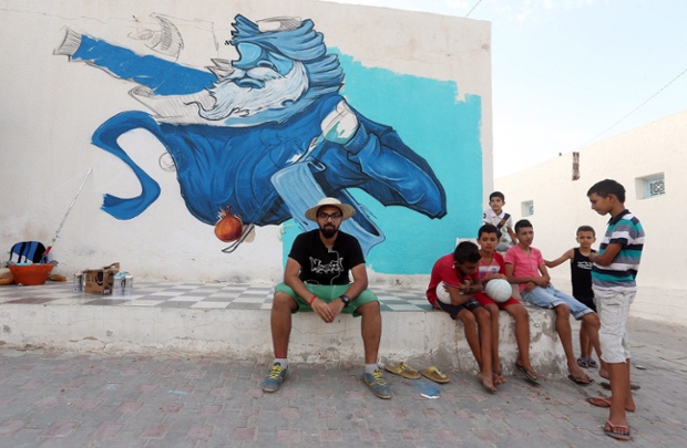 Saudi Arabian artist Deyaa One sit in front of the wall he is decorating as part of the street art project 'Djerbahood'