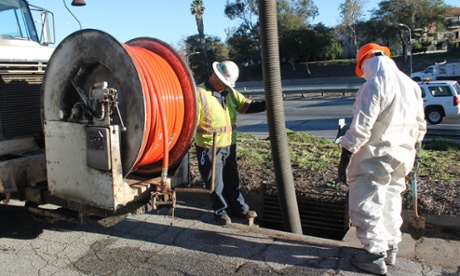 A worker in full hazmat suit oversees the clearing of a roadside drain.