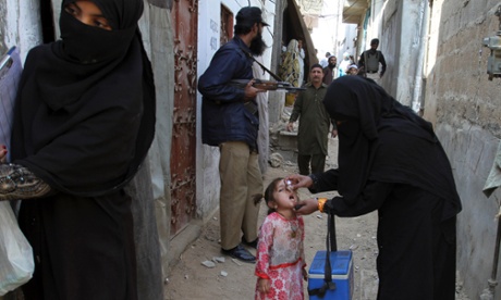 A health worker in Karachi vaccinates a girl against polio while an armed guard provides security.