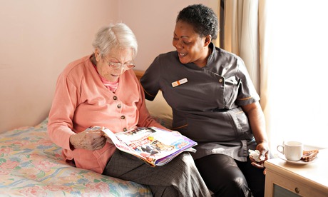 A care-support worker helps an old lady read a magazine on a bed