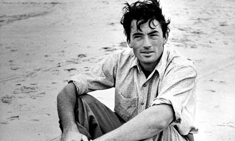A young Gregory Peck sitting on a sandy beach, trousers rolled up, cigarette in hand