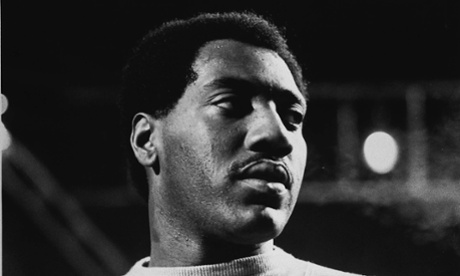 A black and white photo of Otis Redding on stage, the image cropped to his head and shoulders, his head turned slightly so that the stage lighting highlights one side of his face.