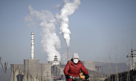 A woman wearing a mask rides past smoking chimneys and cooling towers of a steel plant in Beijing