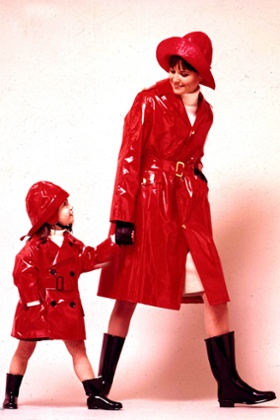 A woman smiling while wearing a red waterproof mac and hat and black wellington boots holds the hand of a young girl wearing a matching outfit, January, 1965, 