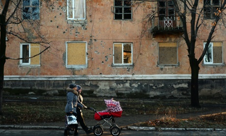 Woman with pram walk past buildings damaged from shells or gunfire in Kommunar 