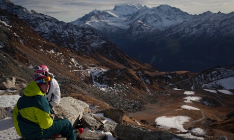 Skiers rest on a mountain overlooking snow-free slopes. Unusually warm, dry weather has ski resorts worldwide on edge.