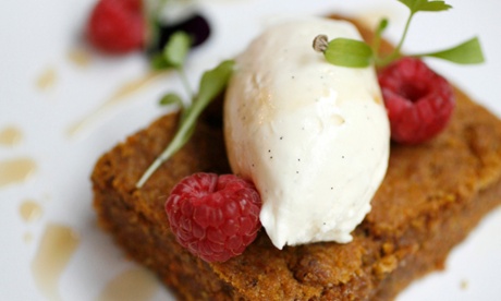 A square of carrot cake next to drops of rum syrup, with Chantilly cream on top and a few raspberrys