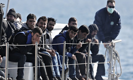 Greek coast guard officer in a mask for fear of infectious disease stands beside a small group of im