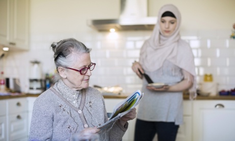 Senior woman reading newspaper with female home caregiver preparing food in kitchen