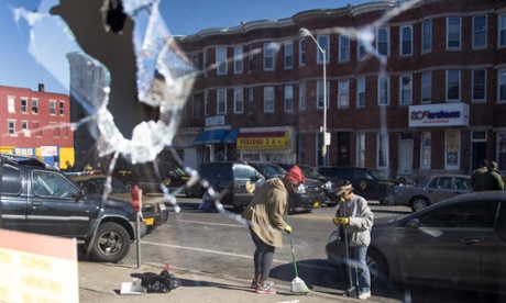 Two women sweeping up the streets are reflected in the broken window of a check-cashing store in Baltimore, Maryland, on April 28.