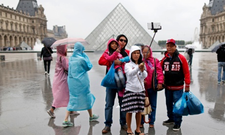 Tourists take a selfie near the Pyramid of the Louvre Museum on a rainy summer day in Paris.