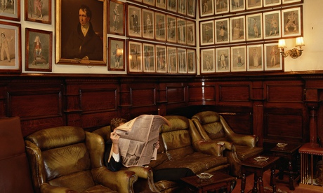 Man behind an open newspaper in an armchair at the Garrick Club, many portraits on the wall