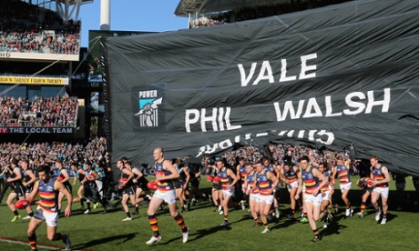 Crows and Power players run through a joint banner to commemorate the late Phil Walsh