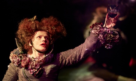 Maxine Peak holds up a wine glass containing red liquid. Her dress appears made from natural materials - faded and brown flowers bursting from the sleeves and neckline.