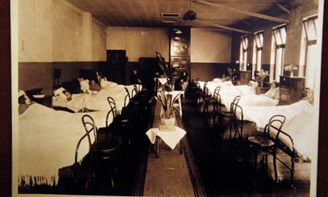 A sepia photograph; two rows of five cast-iron beds, run down the side of a hospital ward, each with a patient (they seem to be women and children). Some beds have tall wooden bedsiders, with vases and flowers.  Bare wood chairs stand at the end of each bed.